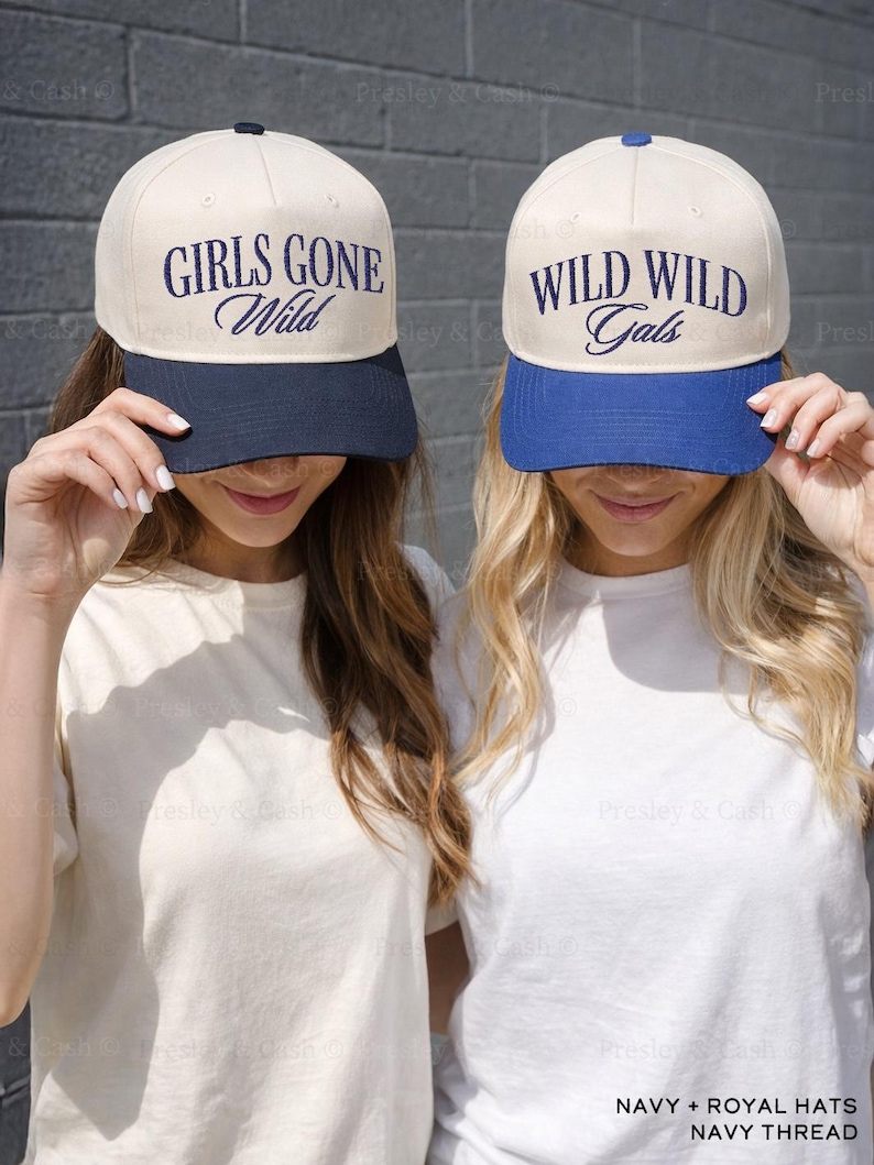 May include: Two baseball caps with contrasting brims. One is navy blue, the other royal blue. The caps have the phrases "GIRLS GONE Wild" and "WILD WILD Gals" embroidered in navy thread. The hats are cream-colored with a dark blue brim.