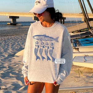 Peut inclure: Un sweat-shirt gris avec un motif de trois sir&egrave;nes tenant un poisson. Le texte "Original designs by PresleyAndCash.com only on Etsy" est imprim&eacute; dans le coin inf&eacute;rieur droit du sweat-shirt.