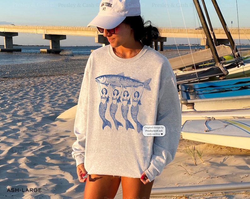 Peut inclure: Un sweat-shirt gris avec un motif de trois sir&egrave;nes tenant un poisson. Le texte "Original designs by PresleyAndCash.com only on Etsy" est imprim&eacute; dans le coin inf&eacute;rieur droit du sweat-shirt.