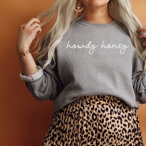 Pode incluir: Uma camisa de moletom cinza com o texto "howdy honey" em escrita branca. A camisa de moletom est&aacute; sendo usada por uma pessoa com cabelo loiro comprido e uma saia de estampa de leopardo.