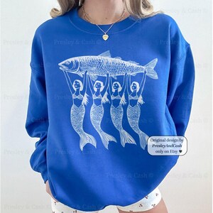 Peut inclure: Un sweat-shirt bleu royal avec un motif blanc de quatre sir&egrave;nes tenant un poisson. Le texte "Original design by PresleyAndCash only on Etsy" est imprim&eacute; dans le coin inf&eacute;rieur droit du sweat-shirt.