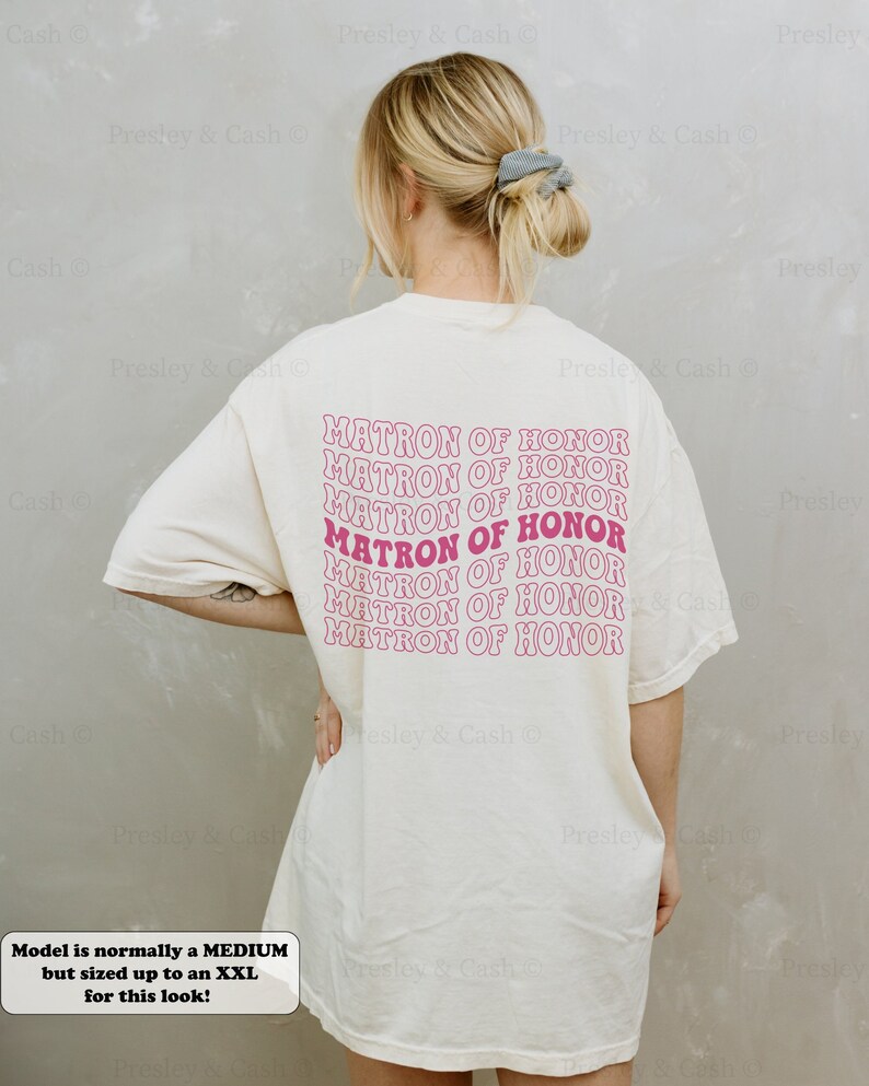 K&ouml;nnte beinhalten: Ein wei&szlig;es T-Shirt mit dem Text "MATRON OF HONOR", der in rosa Schrift auf dem R&uuml;cken wiederholt wird. Das Shirt ist &uuml;berdimensioniert und das Model tr&auml;gt es locker.