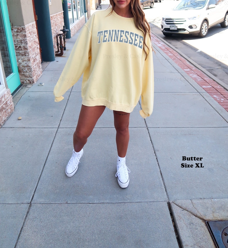 Op de afbeelding: Een geel sweatshirt met het woord "TENNESSEE" in blauw op de voorkant. Het sweatshirt wordt gedragen door een persoon die op een stoep staat. De persoon draagt witte sneakers.