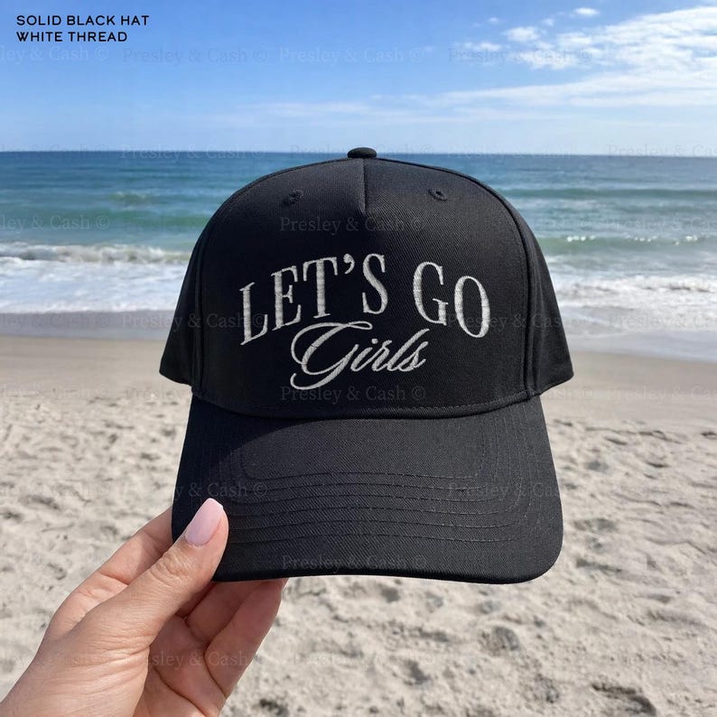 May include: A solid black hat with white thread embroidery. The hat features the text "LET'S GO Girls" in a stylish font. The hat is shown against a beach backdrop with the ocean and sky in the background.