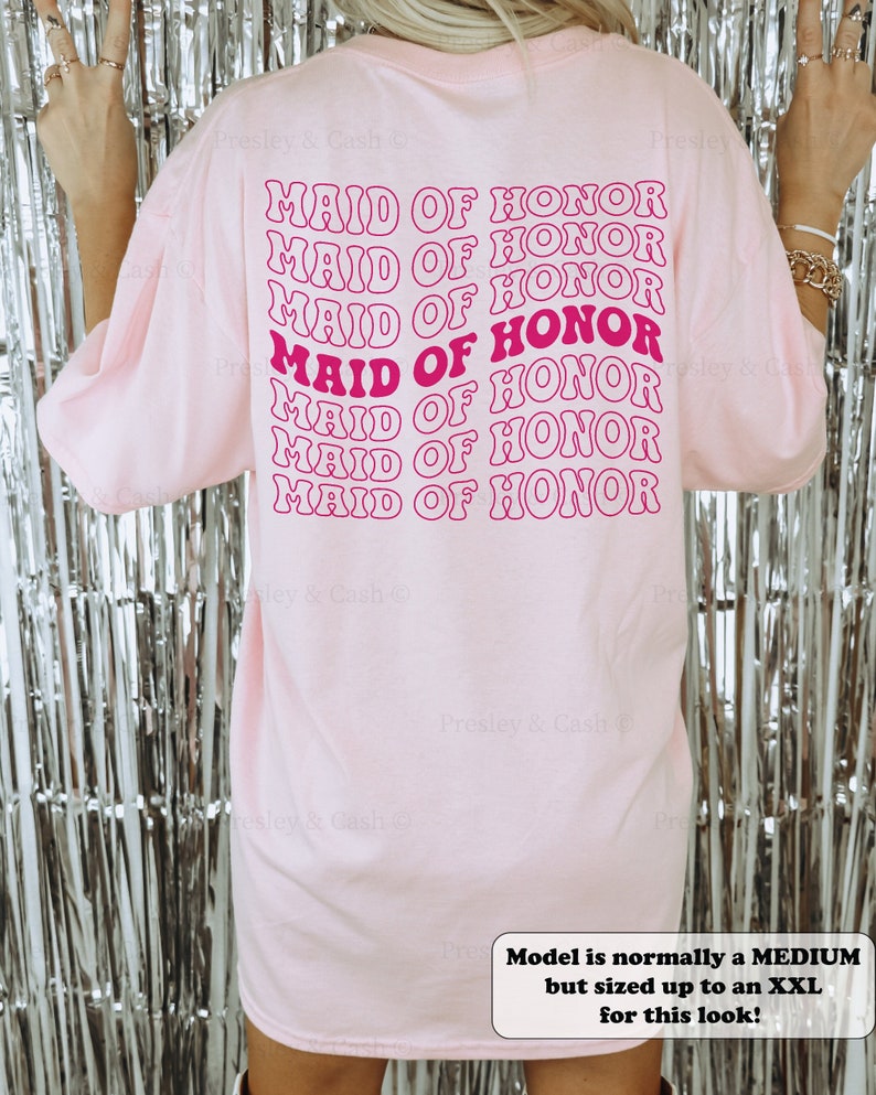 K&ouml;nnte beinhalten: Ein pinkfarbenes T-Shirt mit dem Spruch "Maid of Honor", der in einer Retro-Schriftart in leuchtendem Pink wiederholt wird. Das Model tr&auml;gt Gr&ouml;&szlig;e XXL f&uuml;r eine lockere Passform.