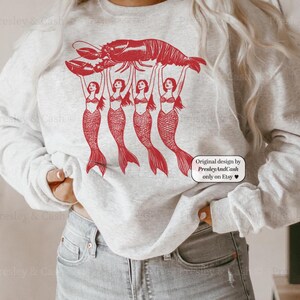 May include: A light gray sweatshirt featuring a red graphic of a lobster held aloft by four mermaids. The design includes the text "Original design by PresleyAndCash only on Etsy".