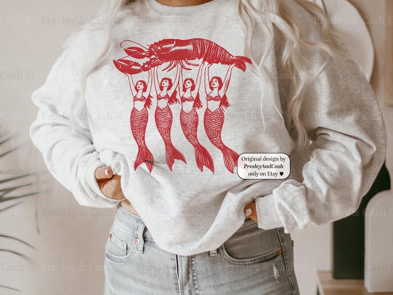 May include: A light gray sweatshirt featuring a red graphic of a lobster held aloft by four mermaids. The design includes the text "Original design by PresleyAndCash only on Etsy".