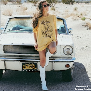 May include: A woman wearing a mustard yellow t-shirt with a vintage graphic of a cowboy riding a horse. The shirt says "Actin' Up" and the woman is standing in front of a white classic car with a license plate that says "California Cowgirl".