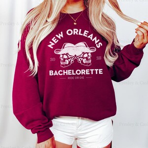 Peut inclure: Sweat-shirt bordeaux avec un imprim&eacute; graphique blanc de deux cr&acirc;nes portant des chapeaux de cow-boy avec le texte "NEW ORLEANS 20 23 BACHELORETTE RIDE OR DIE"