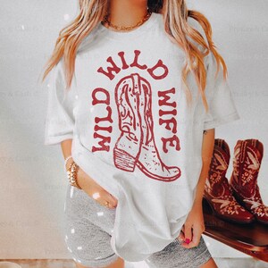 Model wears white t-shirt with “Wild Wild Wife” in dark red text and boot graphic, standing indoors near a pair of cowboy boots. Styled for rustic bridal party or bachelorette trip.