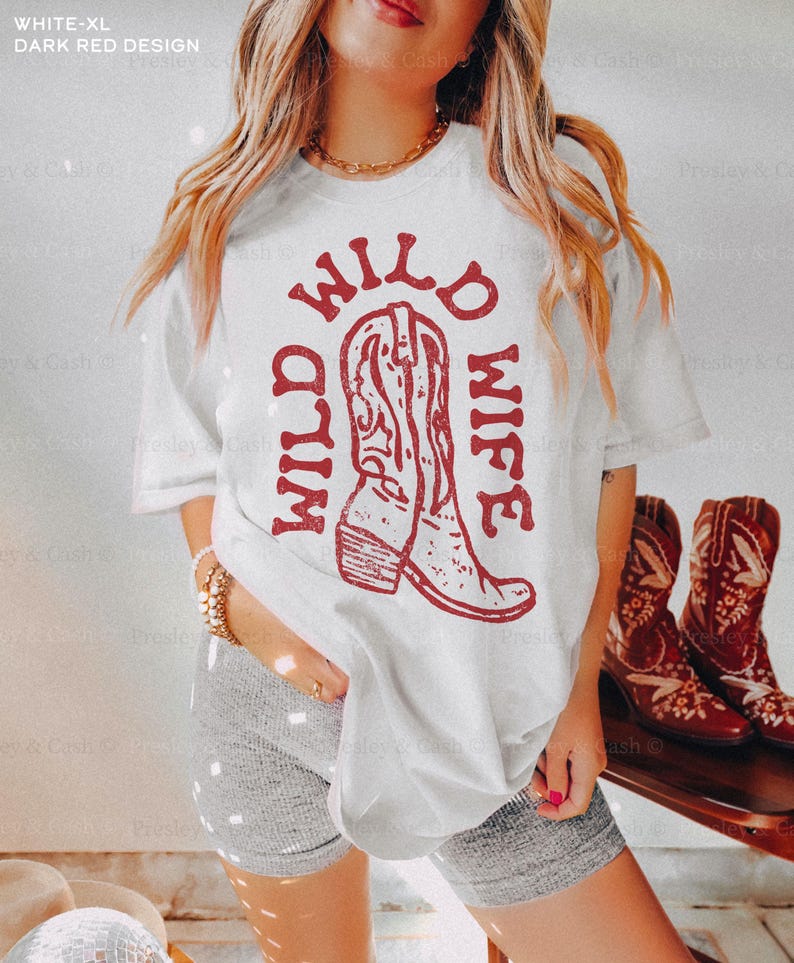 Model wears white t-shirt with “Wild Wild Wife” in dark red text and boot graphic, standing indoors near a pair of cowboy boots. Styled for rustic bridal party or bachelorette trip.