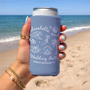 Slim dusty blue can cooler with “Seashells and Wedding Bells” design held on the beach. Features personalized bachelorette text and beach icons like waves, bells, and a cocktail.