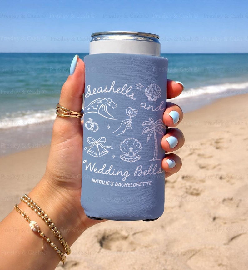 Slim dusty blue can cooler with “Seashells and Wedding Bells” design held on the beach. Features personalized bachelorette text and beach icons like waves, bells, and a cocktail.