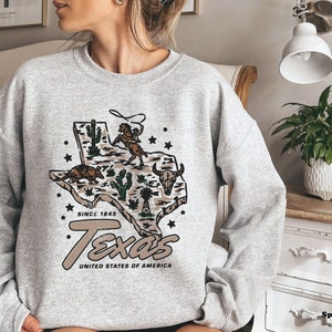 May include: Sport grey sweatshirt featuring a graphic of the state of Texas with a cowboy roping a horse, cacti, and an armadillo. The text "Texas United States of America Since 1845" is printed below the graphic.