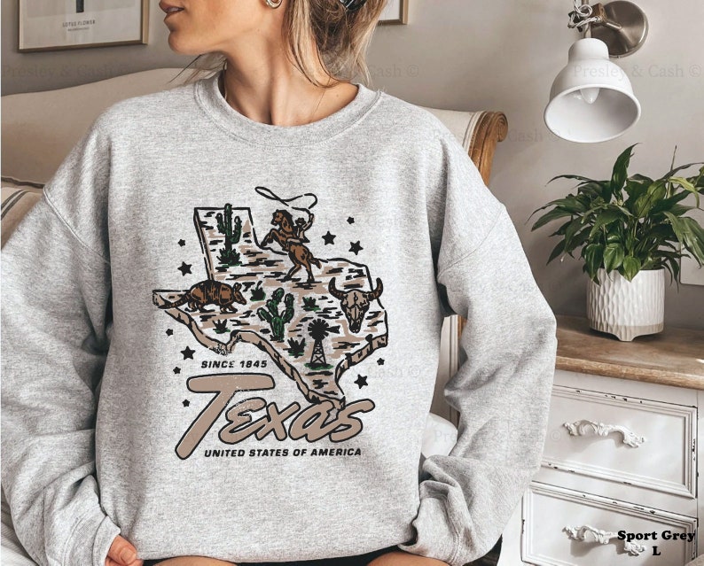 May include: Sport grey sweatshirt featuring a graphic of the state of Texas with a cowboy roping a horse, cacti, and an armadillo. The text "Texas United States of America Since 1845" is printed below the graphic.