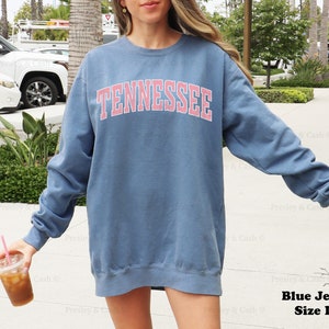 Op de afbeelding: Een blauw sweatshirt met lange mouwen met het woord "TENNESSEE" in roze letters op de voorkant. Het sweatshirt is maat L.