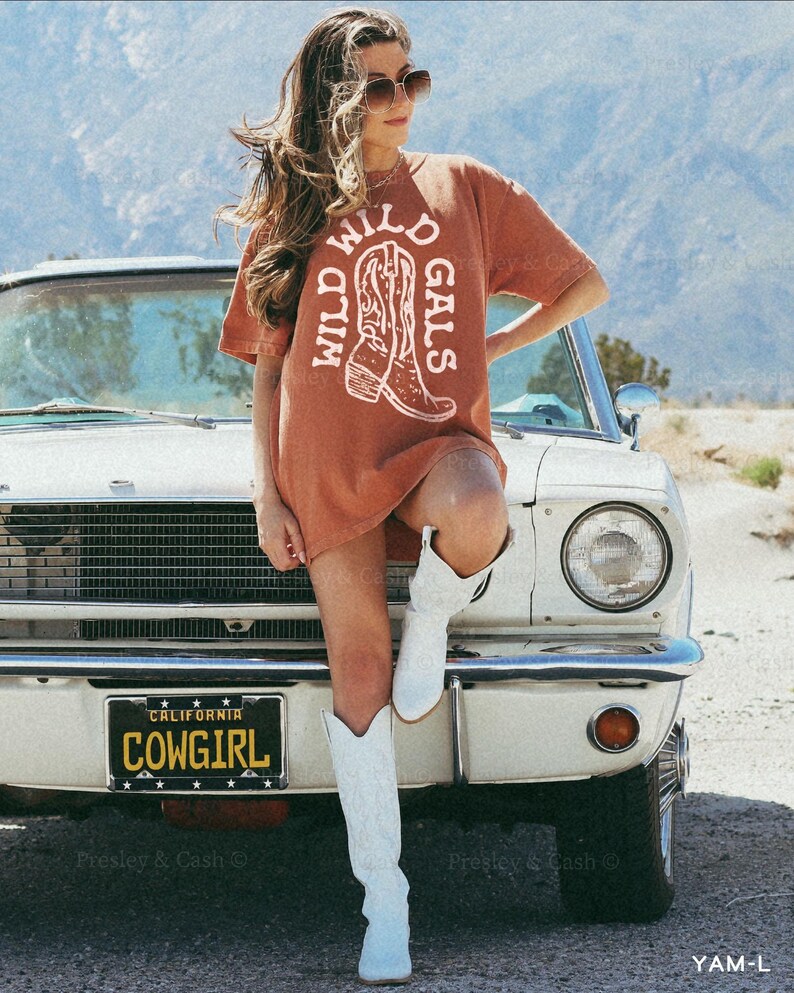 Model in oversized yam-colored shirt with “Wild Wild Gals” in white western font and boot graphic, leaning on vintage car with a “COWGIRL” license plate. Styled for desert western bachelorette trip or Nashville girls weekend.