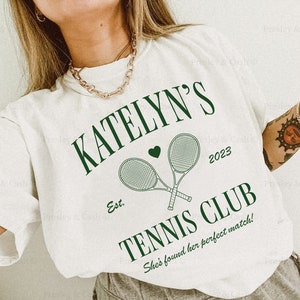 May include: White t-shirt with green text that reads "KATELYN'S Est. 2023 TENNIS CLUB She's found her perfect match!" and a graphic of two tennis rackets with a heart.