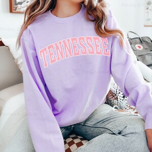 Op de afbeelding: Een lavendelkleurig sweatshirt met het woord "TENNESSEE" in roze op de voorkant. Het sweatshirt wordt gedragen door een persoon en zit op een geruit oppervlak.