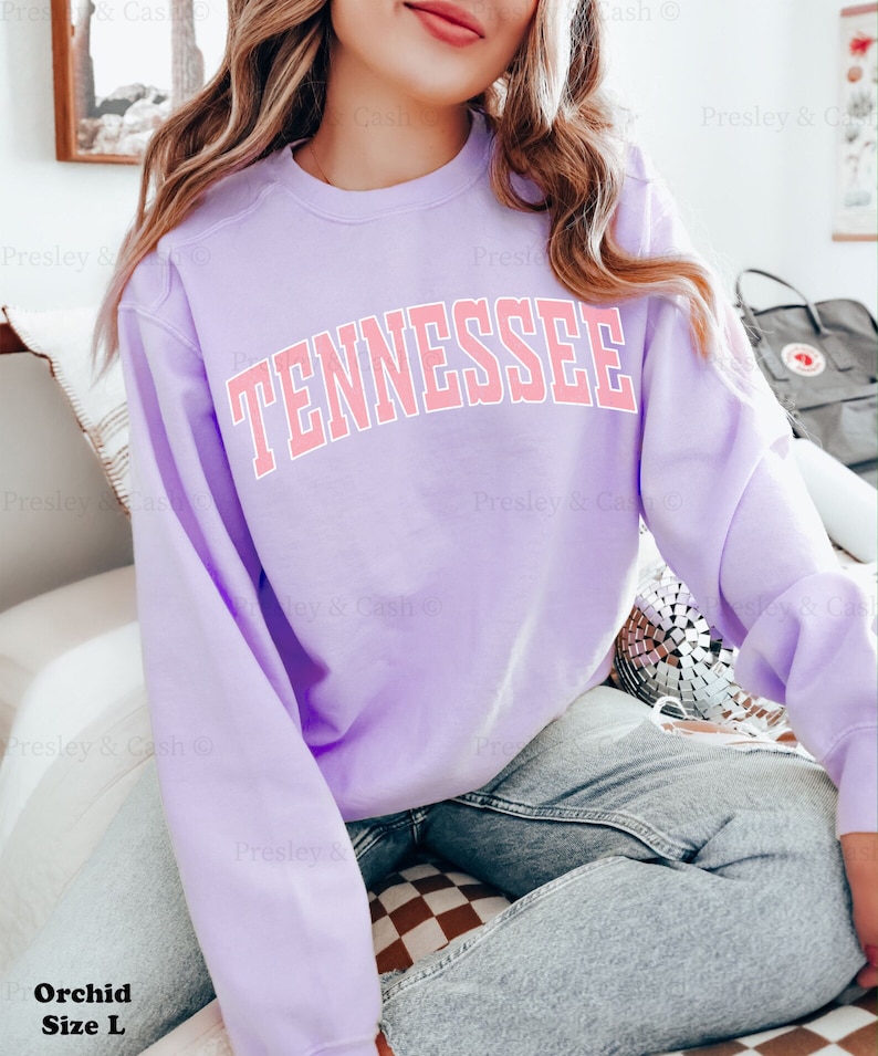 Op de afbeelding: Een lavendelkleurig sweatshirt met het woord "TENNESSEE" in roze op de voorkant. Het sweatshirt wordt gedragen door een persoon en zit op een geruit oppervlak.