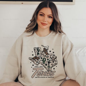 May include: Sand-colored XL sweatshirt featuring a graphic of the Texas state map with cacti, a cowboy, and longhorn. The text "Texas" and "United States of America" are below the map. The sweatshirt has the text "Since 1845".