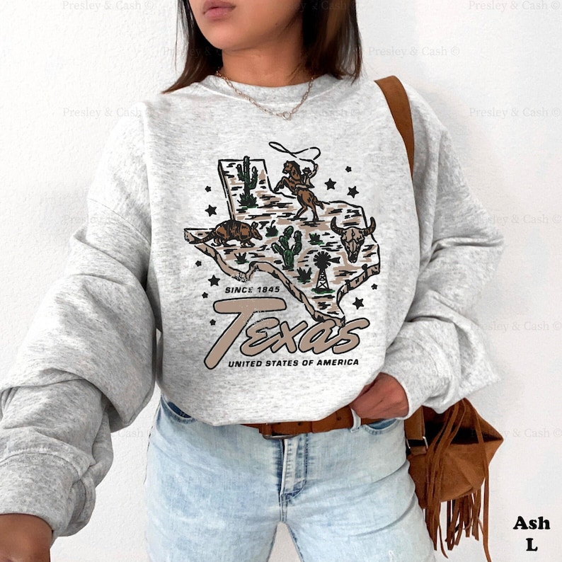 May include: A light grey sweatshirt featuring a detailed graphic of the state of Texas. The design includes a cowboy on a horse, cacti, a windmill, and the text "Texas, United States of America, Since 1845".