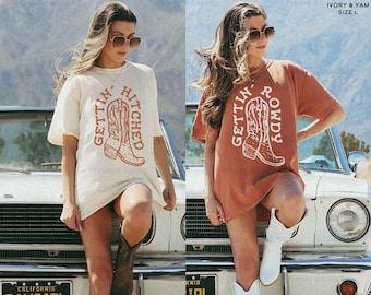 Könnte beinhalten: Elfenbein- und yamfarbene T-Shirts mit den Aufdrucken "GETTIN' HITCHED" und "GETTIN' ROWDY", jeweils mit einer Cowboy-Stiefel-Grafik. Die Shirts sind in Größe L.
