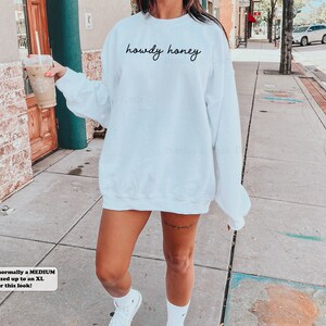 Pode incluir: Camisola branca de tamanho grande com o texto "howdy honey" em escrita preta. A modelo est&aacute; usando a camisola com t&eacute;nis brancos.