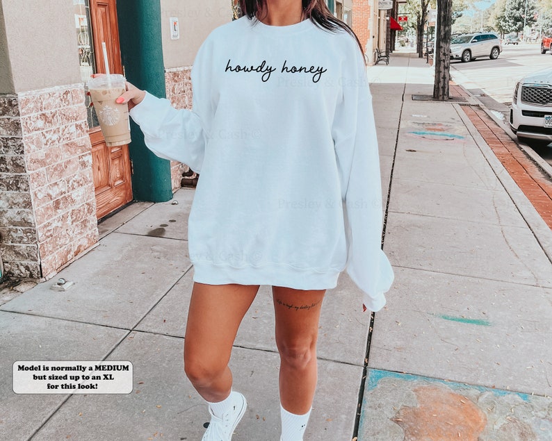 Pode incluir: Camisola branca de tamanho grande com o texto "howdy honey" em escrita preta. A modelo est&aacute; usando a camisola com t&eacute;nis brancos.