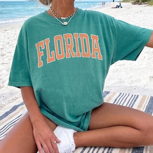 May include: Seafoam green t-shirt with the word "FLORIDA" in orange block letters. The shirt is worn with white sneakers. The image is taken on a beach with a striped blanket. The text "SEAFOAM-XL" is visible in the bottom right corner.