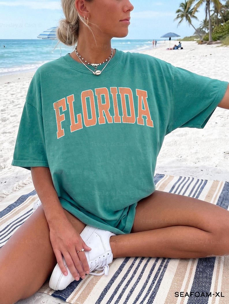 May include: Seafoam green t-shirt with the word "FLORIDA" in orange block letters. The shirt is worn with white sneakers. The image is taken on a beach with a striped blanket. The text "SEAFOAM-XL" is visible in the bottom right corner.