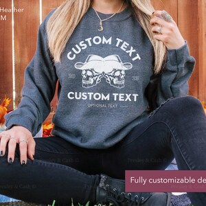 Peut inclure: Sweat-shirt gris chin&eacute; fonc&eacute; avec un motif blanc de deux cr&acirc;nes portant des chapeaux de cowboy. Le texte "CUSTOM TEXT" appara&icirc;t au-dessus et en dessous des cr&acirc;nes. Le texte "OPTIONAL TEXT" appara&icirc;t sous le texte du bas.
