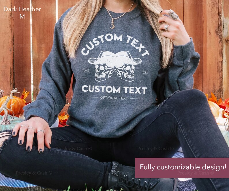 Peut inclure: Sweat-shirt gris chin&eacute; fonc&eacute; avec un motif blanc de deux cr&acirc;nes portant des chapeaux de cowboy. Le texte "CUSTOM TEXT" appara&icirc;t au-dessus et en dessous des cr&acirc;nes. Le texte "OPTIONAL TEXT" appara&icirc;t sous le texte du bas.