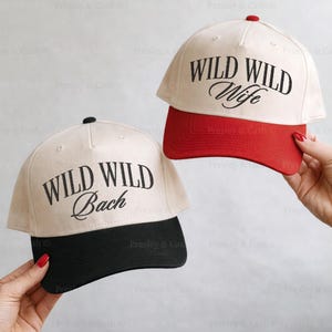 May include: Two baseball caps with the text "WILD WILD Bach" and "WILD WILD Wife" in black script. One cap has a black brim, the other a red brim. Both caps have a cream-colored crown and a black top button.