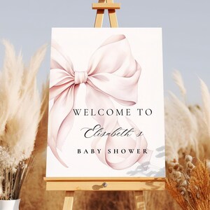 K&ouml;nnte beinhalten: Ein wei&szlig;er Staffelei mit einem Schild, auf dem "Willkommen zu Elizabeths Baby Shower" steht. Das Schild hat eine rosafarbene Aquarell-Schleife. Der Staffelei steht auf einer braunen Oberfl&auml;che mit getrocknetem Pampasgras auf beiden Seiten.
