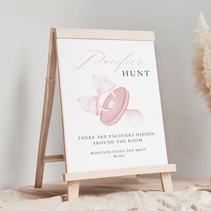 May include: A pink and white sign with the text "Pacifier Hunt" and a pink pacifier with a bow on top. The sign says "There are pacifiers hidden around the room. Whoever finds the most wins!"