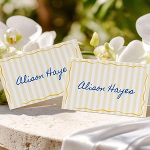Può includere: Due segnaposto a righe gialle e bianche con bordo dorato. Il nome "Alison Haye" è scritto in blu su ogni biglietto. I biglietti sono appoggiati su una superficie di pietra con fiori sullo sfondo.