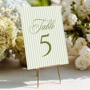 May include: A table number card with a green and white striped pattern. The card reads "Table 5" in a decorative font. The card is displayed on a gold metal stand, perfect for weddings or events.