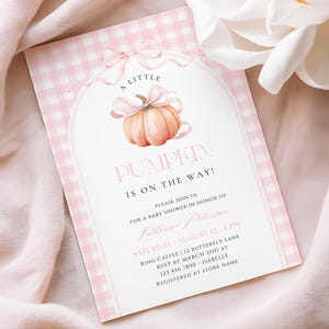 May include: A rectangular baby shower invitation card featuring a light pink and white gingham border. The centre displays an arched frame with a watercolour pumpkin tied with a pink ribbon. Text reads: A LITTLE PUMPKIN IS ON THE WAY!
