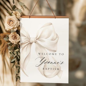 May include: White sign with a large white bow and the text "Welcome to Yvonne's Baptism" in black lettering.
