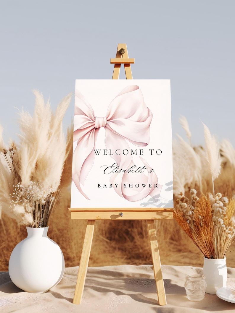 K&ouml;nnte beinhalten: Ein wei&szlig;er Staffelei mit einem Schild, auf dem "Willkommen zu Elizabeths Baby Shower" steht. Das Schild hat eine rosafarbene Aquarell-Schleife. Der Staffelei steht auf einer braunen Oberfl&auml;che mit getrocknetem Pampasgras auf beiden Seiten.