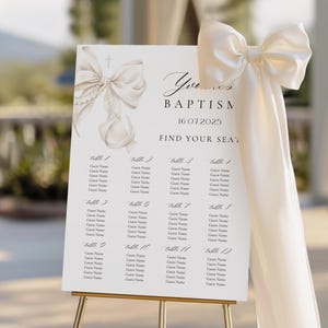 May include: A white baptism seating chart on a gold easel. The chart features a decorative bow and the text "Yvonne's Baptism 16.07.2025 Find Your Seat." It includes space for guest names, organized by table number. A cream-colored bow adorns the side.