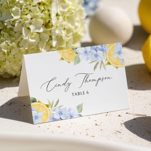 Può includere: Un segnaposto bianco piegato con illustrazioni ad acquerello di limoni, fiori blu e verde. La carta presenta il nome "Cindy Thompson" in elegante corsivo e "TABLE 6" in un carattere più piccolo.