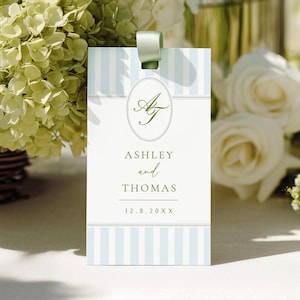 Könnte beinhalten: Eine rechteckige Hochzeitsplatzkarte mit hellblauen und weißen Streifen, mit den Namen "Ashley and Thomas" und dem Datum "12.8.20XX". Die Karte hat ein grünes Band und ein grünes Monogramm in einem Oval.