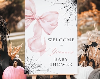 May include: A baby shower welcome sign on a wooden easel. The sign features a large pink bow, spiderwebs, and spiders. Text reads "Welcome to Yvonne's Baby Shower."