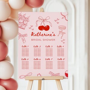 May include: A pink bridal shower seating plan displayed on an easel. The chart has the text "Katherine's Bridal Shower" with illustrations of cherries, bows, and cupcakes. It lists table numbers and space for guest names.