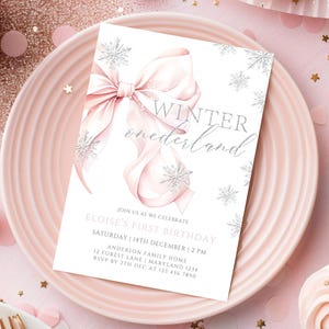 May include: A white invitation card with a pink bow and silver snowflakes. The text on the card reads "Winter onederland" and includes details for Eloise's first birthday party.