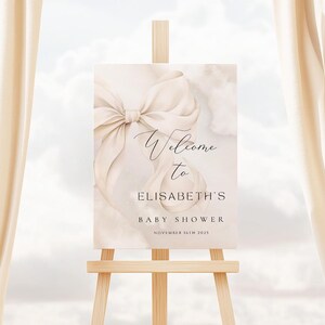 May include: Welcome to Elisabeth's Baby Shower sign with a large white satin bow on a watercolor background. The sign is on a wooden easel.