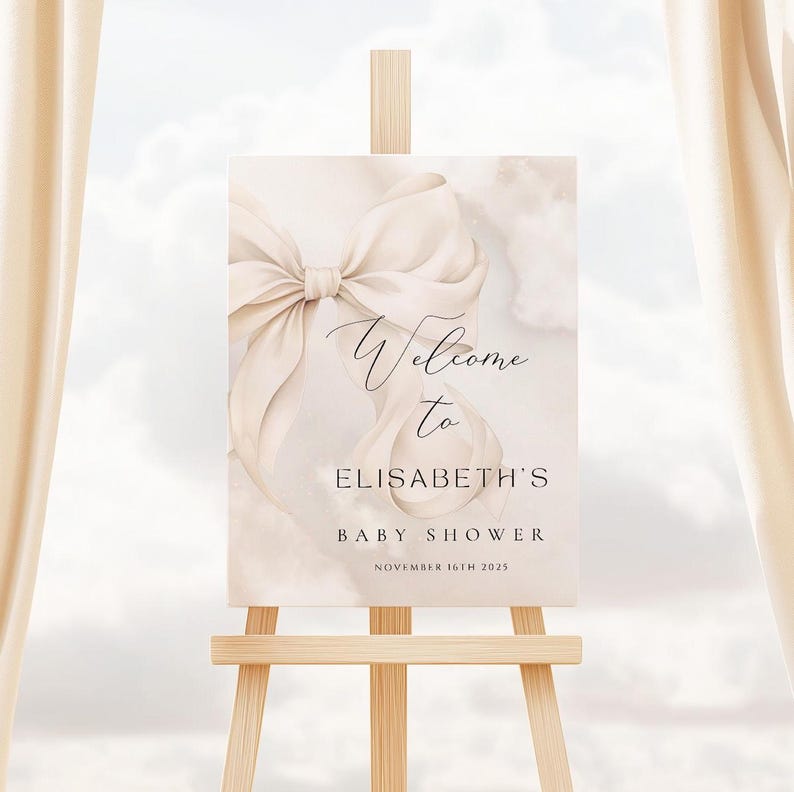 May include: Welcome to Elisabeth's Baby Shower sign with a large white satin bow on a watercolor background. The sign is on a wooden easel.