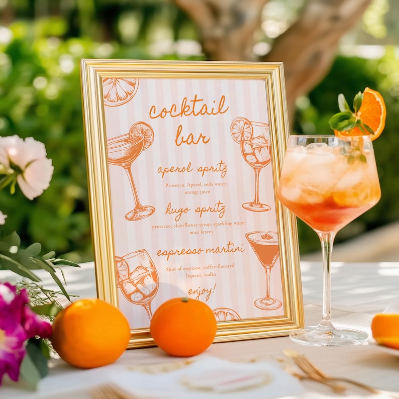 May include: A gold-framed sign with the words "cocktail bar" and drink recipes, including Aperol Spritz and Espresso Martini. A cocktail glass with an orange slice garnish sits next to the sign, along with two oranges.